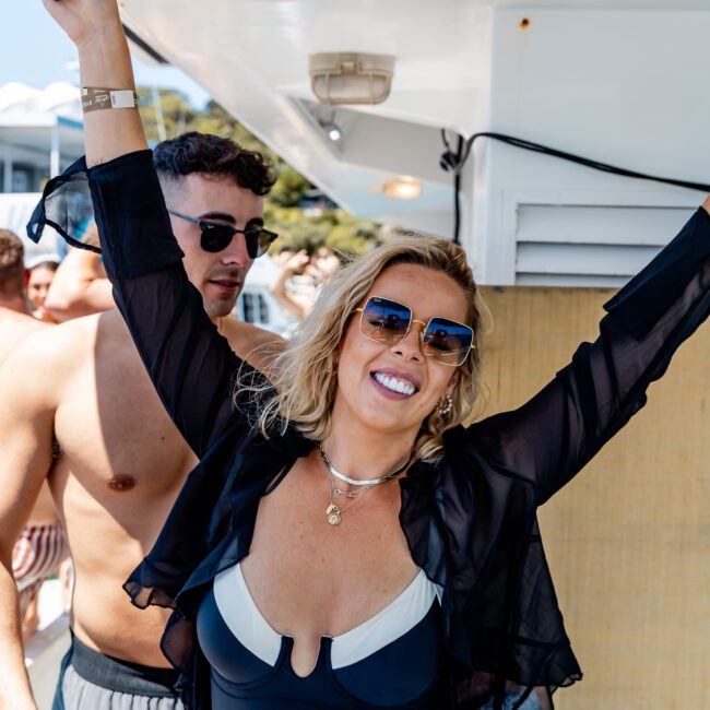 A woman in a black swimsuit and sunglasses smiles with her arms raised on a yacht. A man in sunglasses stands nearby. The background shows other people and a sunny outdoor setting.