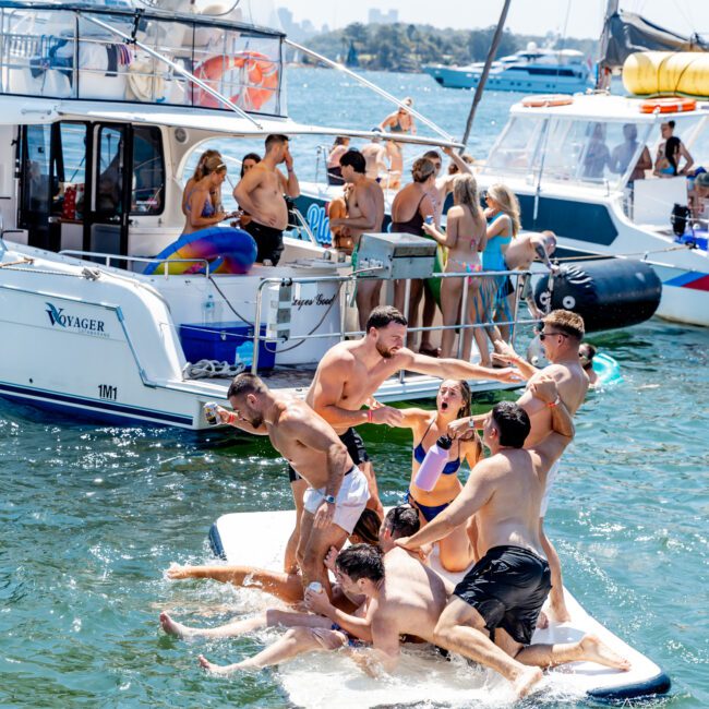 A group of people having fun on a floating mat in the water near a yacht. Some are playfully pushing each other into the water, while others laugh and watch. The scene is lively, with more people on the boat in the background.