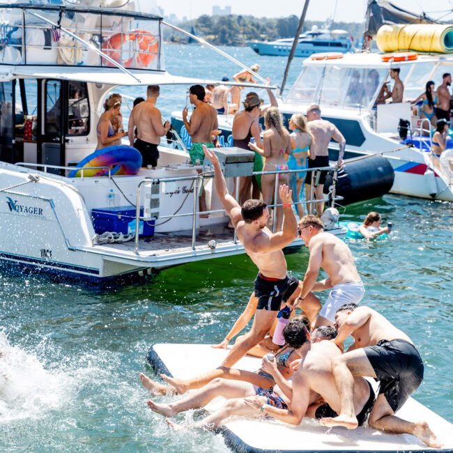 A group of people having fun on a floating mat in the water near a yacht. The scene is lively, with several individuals on the boat and others in the water enjoying the sunny day.