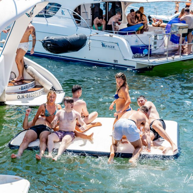 A group of people enjoying the sun on inflatable mats and boats in a lively water setting. Some are sitting, while others are dipping their feet in the water. A few boats are nearby, and the atmosphere is relaxed and joyful.