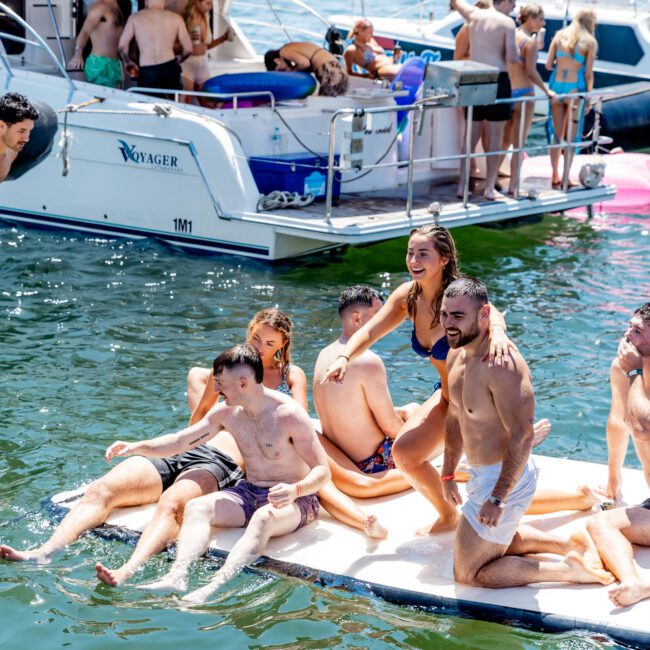 A group of people enjoy a sunny day on a lake, sitting and standing on a floating platform next to a boat named "Voyager." In the background, more individuals socialize on the boat and nearby.