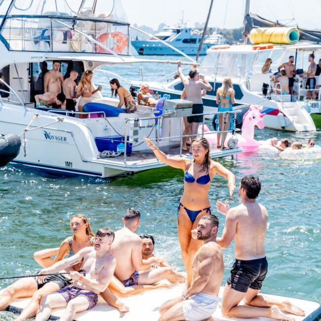 People enjoying a sunny day on a boat party. A group in swimwear relaxes on a floating platform in the water. Boats and people swimming are visible in the background, alongside an inflatable pink flamingo.