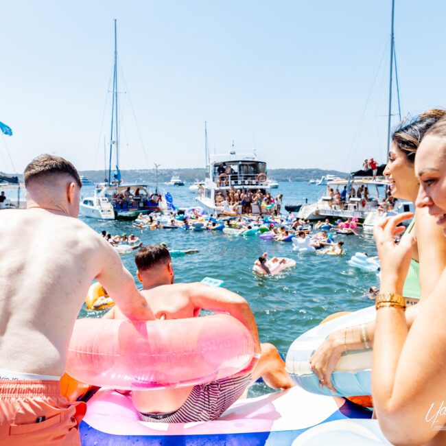 People enjoying a sunny day on the water, lounging on colorful inflatables. Boats and yachts are anchored nearby, and individuals are socializing and relaxing. The scene conveys a lively and festive atmosphere on the open water.