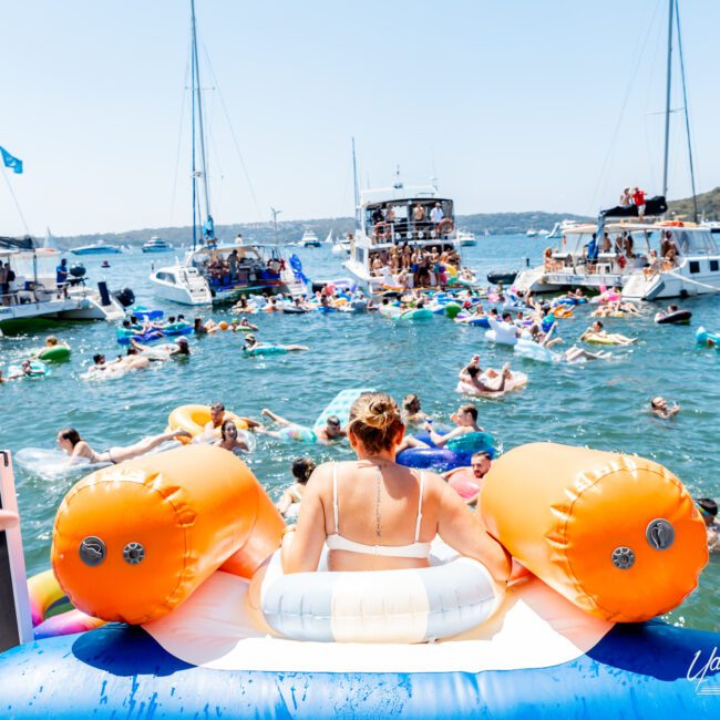 A vibrant scene at a yacht social event on the water. A woman sits on an inflatable near a dock, surrounded by people swimming and floating on inflatables. Boats and yachts are anchored in the background under a clear sky.