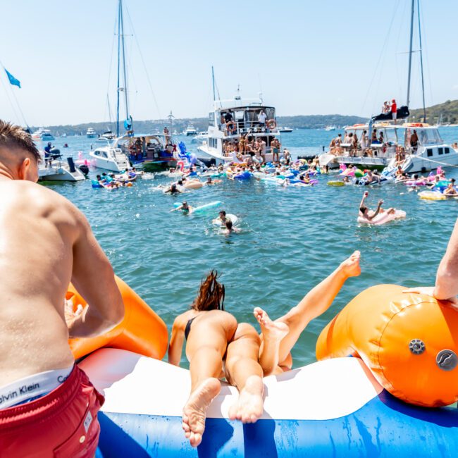 A lively boat party scene with people swimming and lounging on floats in a sunny bay. A woman is sliding into the water from a boat, while others gather around enjoying the festive summer atmosphere. Multiple boats are anchored nearby.