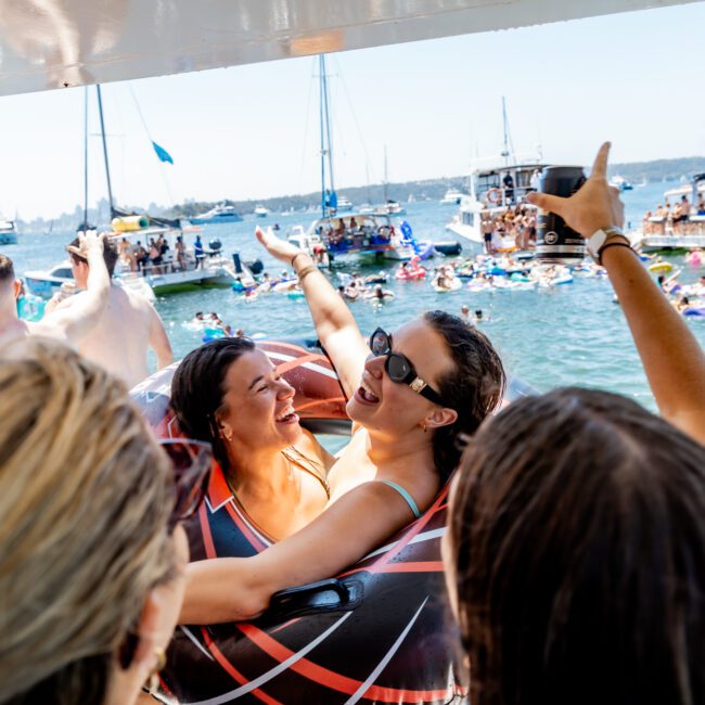 A group of people enjoy a sunny day on the water. Two women in swimsuits, wearing sunglasses and laughing, embrace on a boat. Several boats and people swimming in the background. The atmosphere is lively and festive.