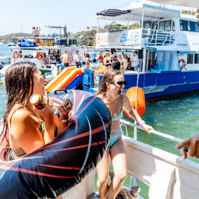 Two women laughing on a boat, one holding a black inflatable. They're surrounded by water and other boats, with people enjoying a sunny day on the water.