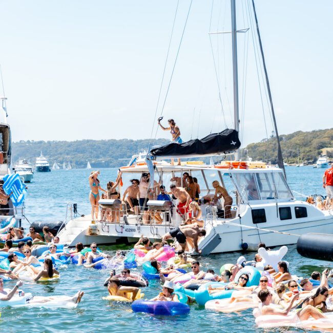 A vibrant gathering of people on a boat and in the water, lounging on inflatables. They are enjoying a sunny day with yachts and scenic hills in the background. Summer fun and relaxation are in full swing.