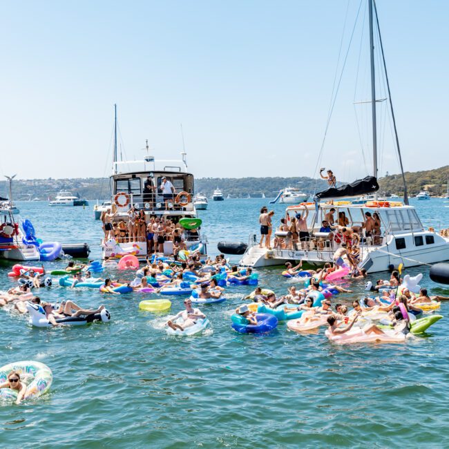 A lively scene on a sunny day with a group of people enjoying a boat party. Several boats are anchored close together, surrounded by people floating on colorful inflatables in the clear blue water under a bright sky.