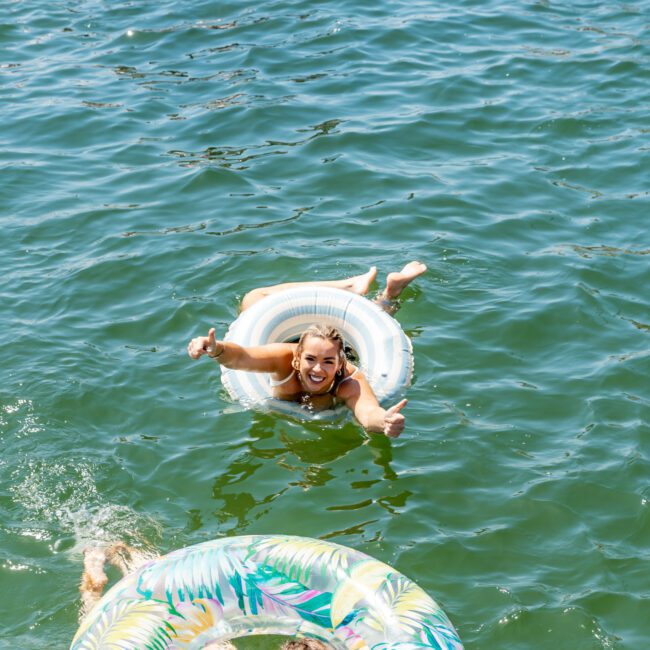 People enjoying a sunny day floating on inflatable rings in a lake. The water is clear, and various colorful floats can be seen around. One person in the foreground is smiling and giving a thumbs-up.