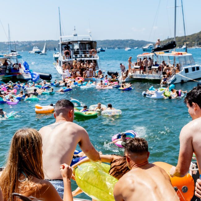 A crowded boat party scene on a sunny day shows people enjoying the water on inflatable floats. Boats are anchored around, and participants in swimsuits are socializing and swimming in the vibrant blue sea.