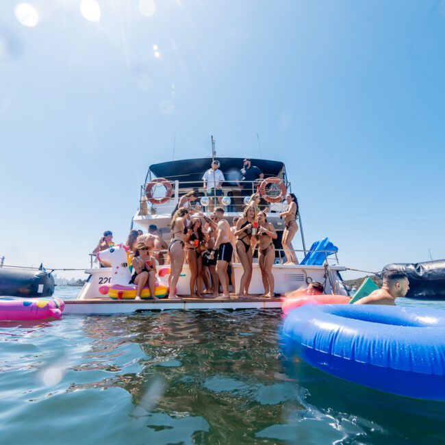 A group of people enjoying a sunny day on the back of a yacht, surrounded by colorful inflatable floats in the water. The scene is lively, with people chatting and relaxing under a clear blue sky.