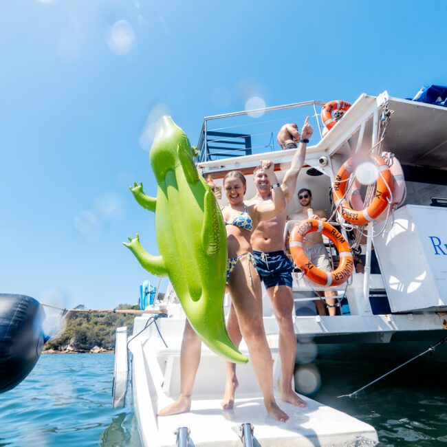 Three people in swimwear on a boat with a "Rum Runner Cruise" sign. One holds a large inflatable green dinosaur. The boat is docked near scenic water. Bright, sunny day with water droplets on the camera lens.