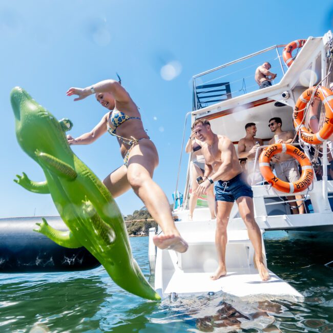 A person in a swimsuit jumps off a boat onto a green inflatable crocodile in the water. Other people watch from the boat, with the sky clear and sunny. Life rings are visible on the boat's railing.