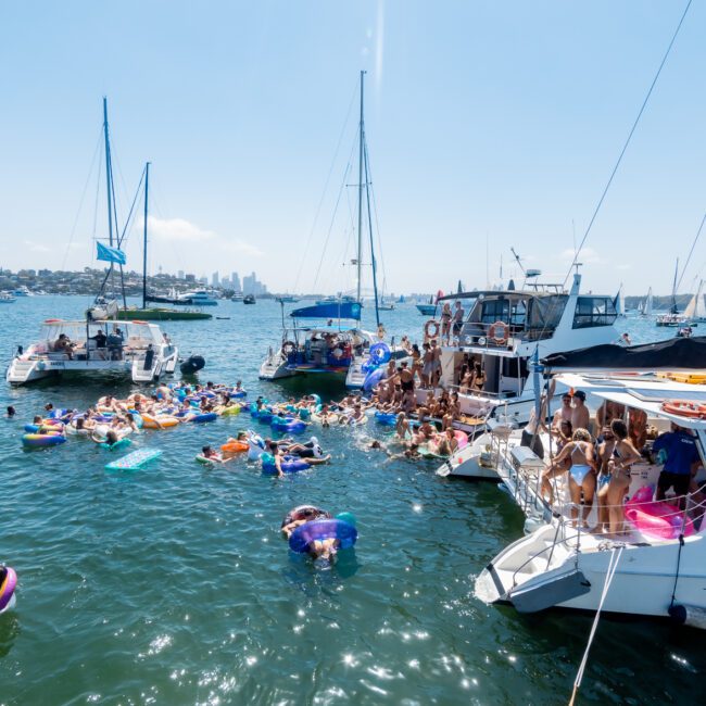 A lively scene on the water with people enjoying a yacht party. Several boats are anchored close together, with many individuals swimming and floating on inflatables. The background shows a city skyline under a clear blue sky.