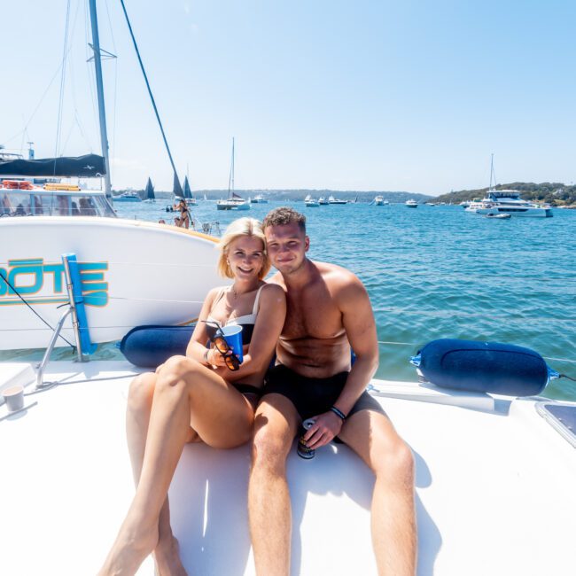 A couple sits on the deck of a boat, smiling and holding drinks. The woman is wearing a bikini, and the man is shirtless. Other boats are visible on the blue water under a clear sky.