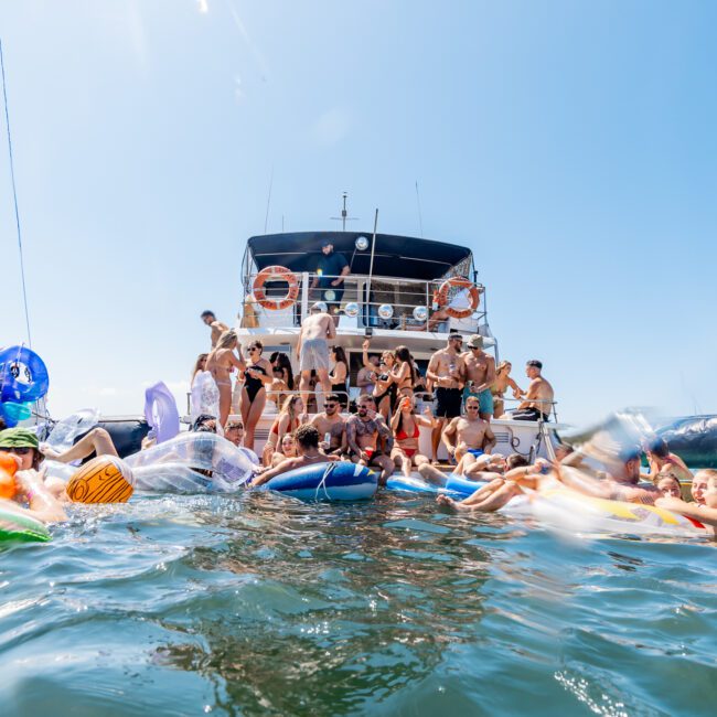 A large group of people enjoying a sunny day on a yacht and in the water, with inflatable floats surrounding them. The yacht is anchored, and there’s a joyful atmosphere under a clear blue sky.