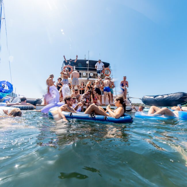 A lively gathering on a yacht with people swimming, lounging on inflatables, and enjoying the sunny day. The yacht is anchored in clear blue water, and a group is gathered on the deck. Various floaties are seen, including a unicorn.