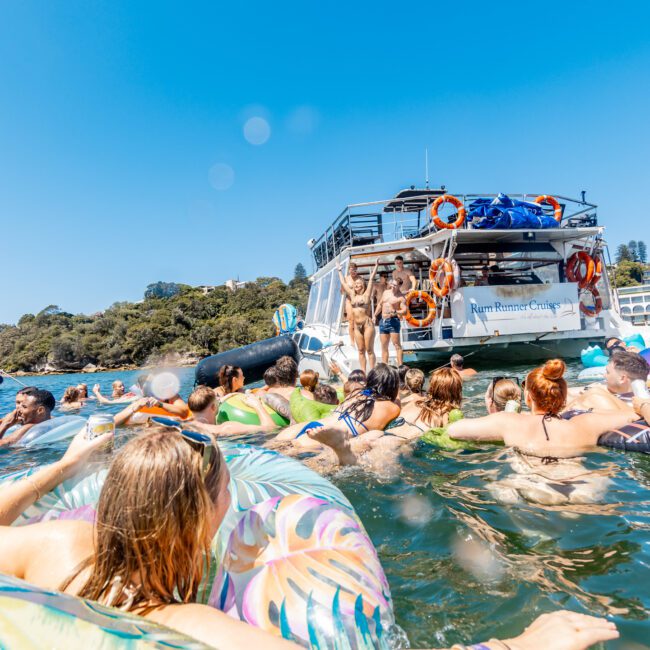 A group of people relax in the water on colorful inflatables near boats on a sunny day. Several people are on a boat with a "Yacht Social Club" sign. The background features trees and a clear blue sky.