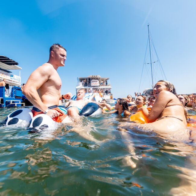 A group of people enjoying a sunny day in the water near boats. One person is on a panda-shaped float, and others are swimming and socializing. The sky is clear, and the atmosphere is lively and fun.