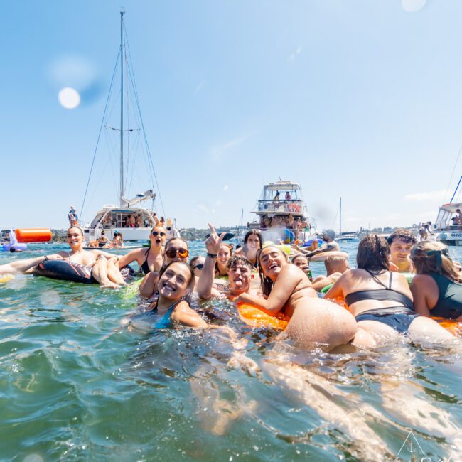 A group of people enjoying a sunny day in the water, surrounded by inflatable floaties. Sailboats are visible in the background. The scene depicts a fun and lively atmosphere, with clear blue skies and several people posing for the camera.