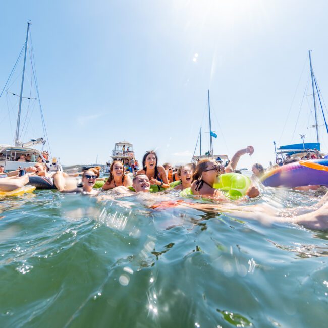 A group of people joyfully floating in the ocean with inflatable tubes. Several boats are anchored in the background under a clear blue sky. The water is bright and lively, reflecting the sunny atmosphere.