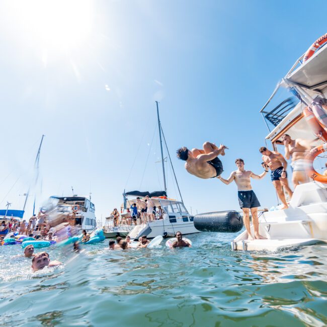 People enjoying a sunny day on the water; some are on boats while others swim. A person performs a backflip into the sea from a boat. Other boats and people can be seen in the background under a clear blue sky.