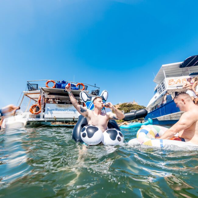 People are enjoying a sunny day on the water, floating on inflatables near boats. One man is on an inflatable panda, and others are in life vests and swimsuits, relaxing under the clear blue sky.