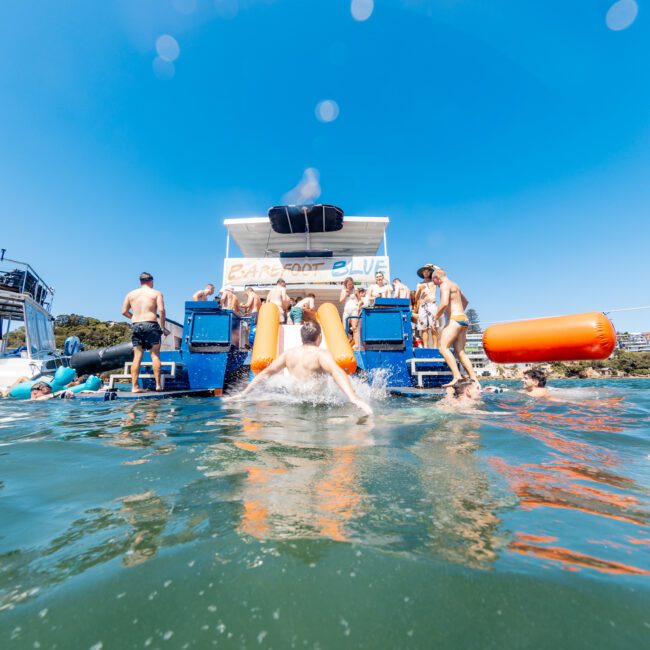 A group of people enjoy a sunny day on the water near a party boat with slides. Two individuals are seen sliding into the water, surrounded by others swimming and relaxing. The backdrop features trees and clear blue skies.