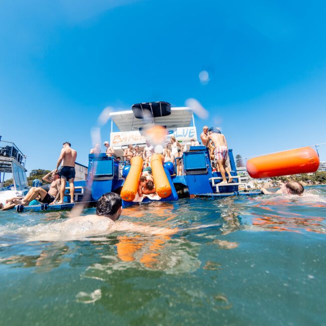 People are enjoying swimming and relaxing around a boat in clear blue water. One person is in an inflatable orange tube near the boat's platform. The sky is clear and sunny. The boat has a banner that reads "Birthday Cruise.