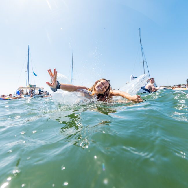 A woman joyfully splashes in the water, surrounded by people and yachts. The sun is shining brightly on a clear day, and sailboats are visible in the background.