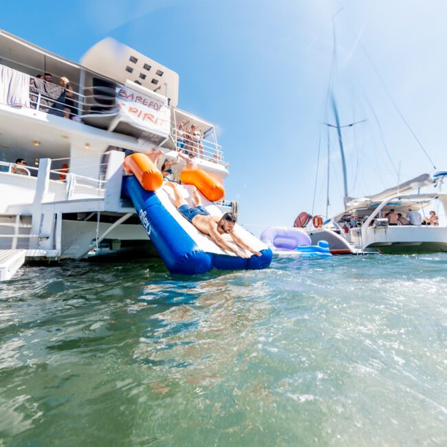 A person slides down a colorful inflatable slide attached to a boat into the water. Other boats and people can be seen in the background under a clear blue sky.