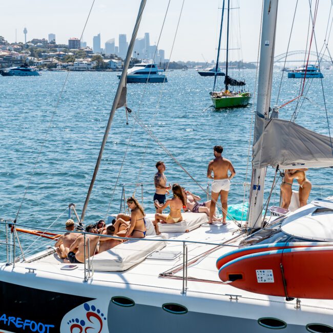 A group of people relaxing on the deck of a sailboat, enjoying sunny weather. They are anchored in a bay filled with other boats. A city skyline and a bridge are visible in the background under a clear blue sky.