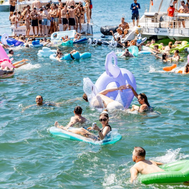 A lively scene of people enjoying a sunny day on a lake with colorful inflatable floats. Several boats are anchored, and groups of people are relaxing on deck and in the water. Hills and trees are visible in the background.