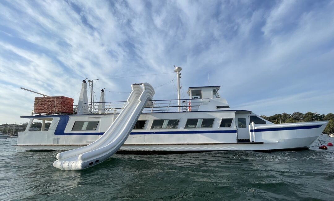 A white and blue boat with two curved slides on its side is anchored on calm water. The sky is partly cloudy, and there are trees in the background.