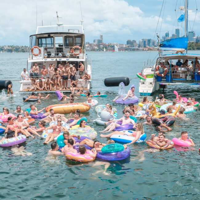 A lively group of people floats on colorful inflatables in a harbor, surrounded by two boats. The skyline is visible in the background under a partly cloudy sky. The scene is festive and summery.