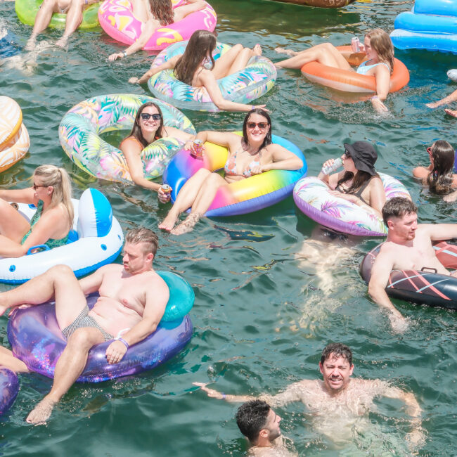 A crowded scene of people floating on colorful inflatable tubes in the water, enjoying a sunny day. The group includes men and women of varying ages, some wearing sunglasses and smiling. Inflatable shapes include donuts and animals.