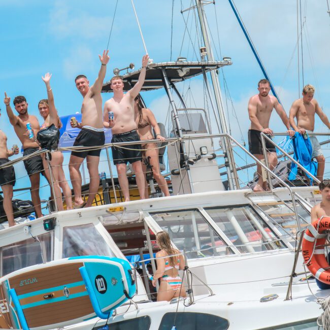 A group of people stands on a boat in swimsuits, smiling and waving at the camera. Paddleboards are visible, and the sky is clear and blue. Everyone appears to be enjoying a sunny day on the water.