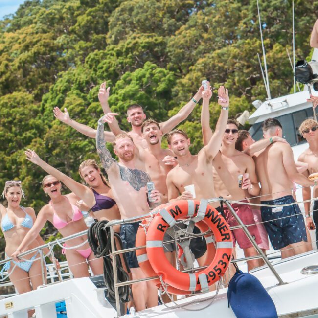 A group of people in swimwear are joyfully posing on a boat, surrounded by lush greenery. They are smiling, waving, and holding drinks, suggesting a festive and relaxed atmosphere.