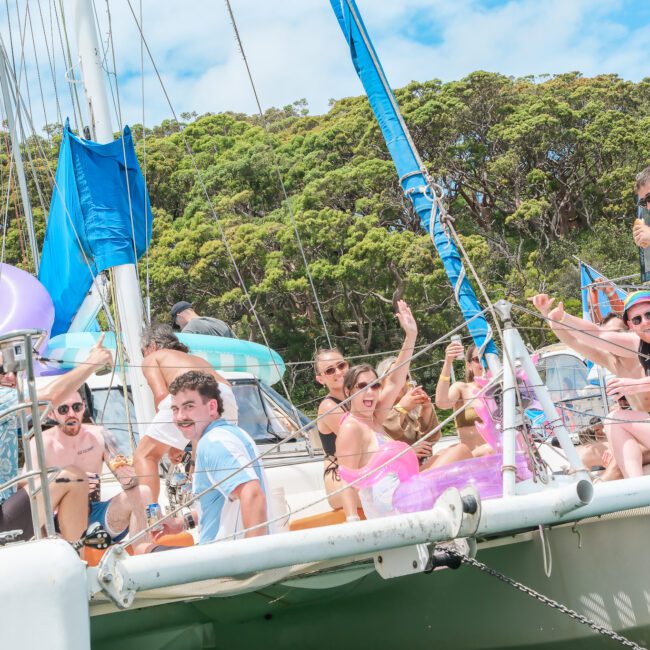 A group of people on a sailboat are enjoying a sunny day. They are sitting and standing on the deck, surrounded by inflatable pool accessories. Some are waving and holding drinks. The sea and a forested shoreline are visible in the background.