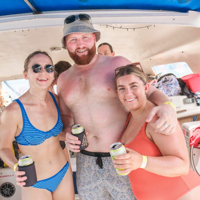 Three people smiling on a boat, enjoying drinks. They are casually dressed in swimwear, with one person wearing a bucket hat. The background shows boat equipment and a canopy providing shade. It's a sunny day, suggesting a fun, relaxed outing.