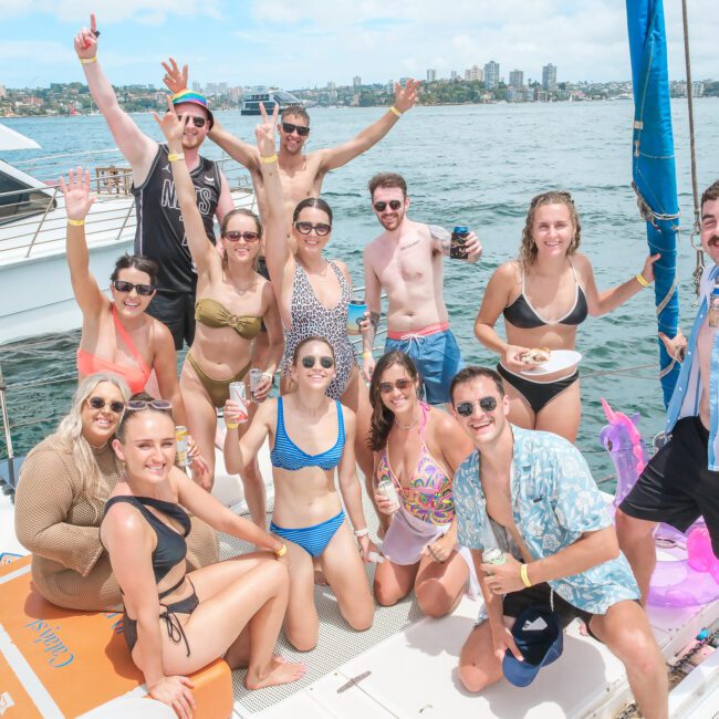 A group of people in swimwear enjoying a sunny day on a boat. They are smiling, holding drinks, and posing for the camera. The background features water and a distant city skyline.