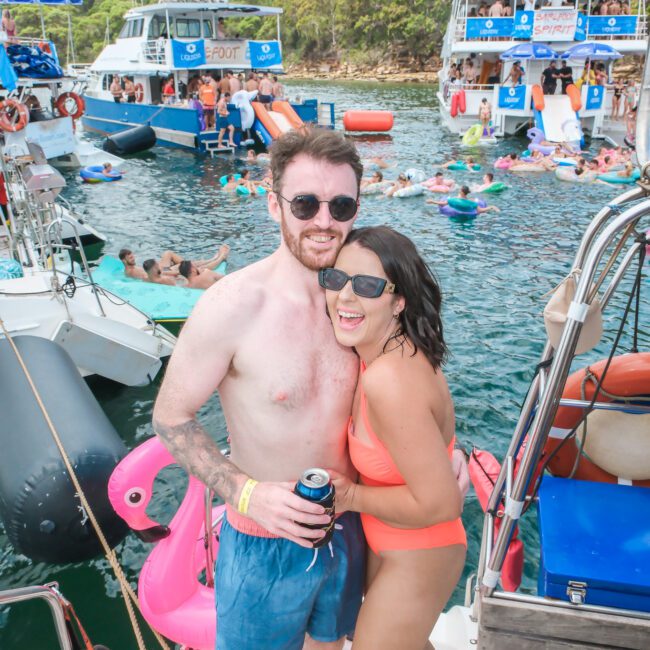 A smiling couple wearing swimsuits pose together on a boat. They are surrounded by other boats and people enjoying the water. Inflatable pool floats and a party atmosphere are visible in the background.