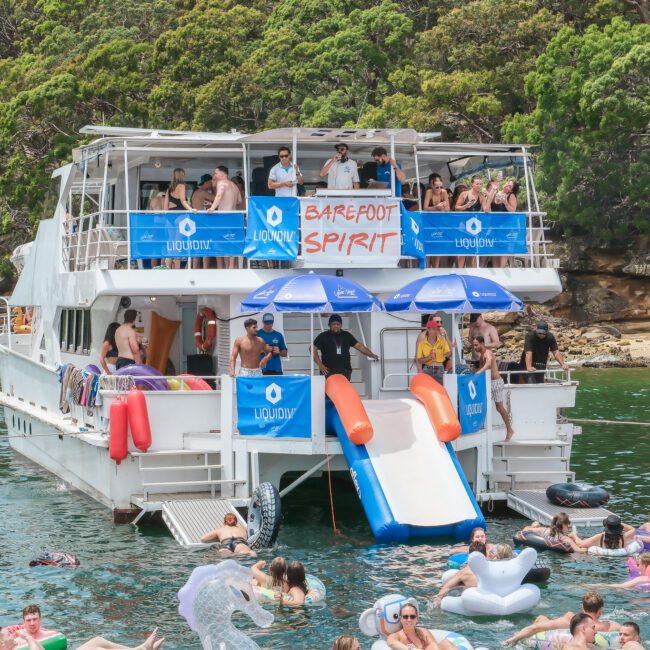 A lively boat party with people enjoying themselves on a large double-decker boat. A banner reads "Barefoot Spirit." Inflatable slides lead into the water, where more people relax on floats. The boat is surrounded by greenery and calm water.