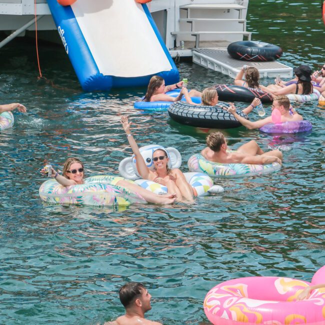 A lively scene of people enjoying a sunny day on a lake, floating on colorful inflatable rings and lounging by a waterslide.