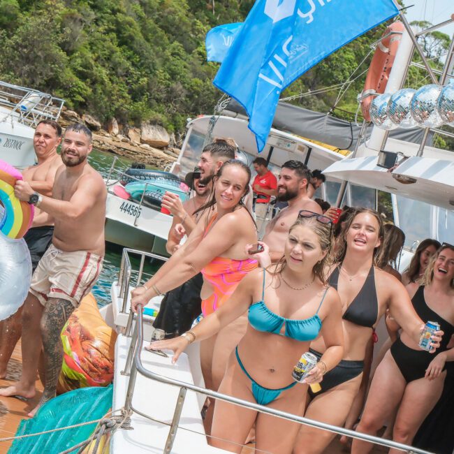 A group of people in swimsuits enjoy a party on a boat. Some are holding drinks and laughing. A rainbow float is in the foreground, with greenery and other boats in the background. The scene is lively and festive.