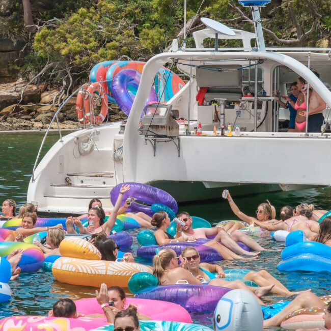 A group of people relaxing on colorful inflatable floats in the water near a boat. The scene is lively and festive, set against a backdrop of green foliage and clear blue water. The boat is anchored and several floats are clustered nearby.