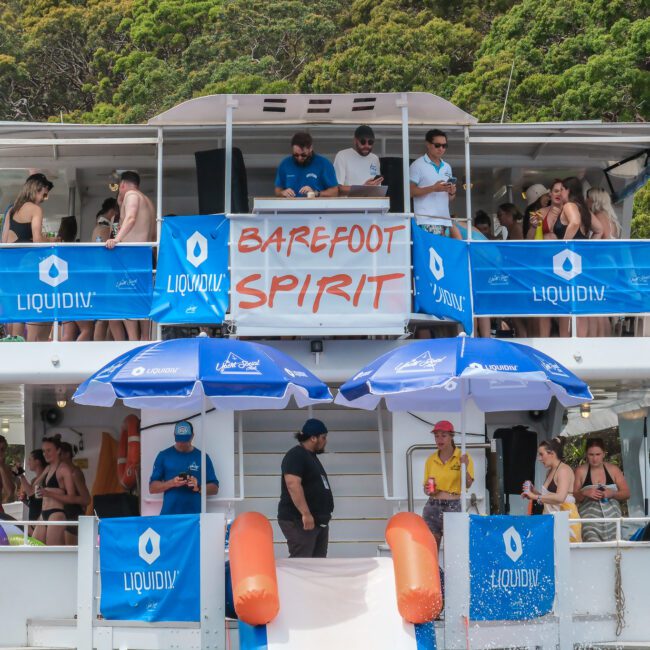 A large boat with two decks is crowded with people. Several individuals are standing and socializing under a "Barefoot Spirit" banner. Blue umbrellas and banners from "LiquidIV" brand are displayed. The background features lush greenery.