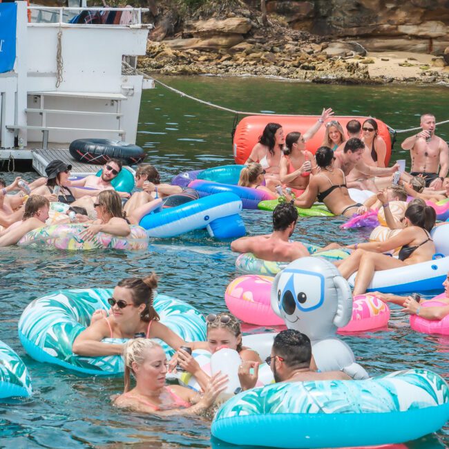 A vibrant scene of people relaxing and socializing on colorful inflatable floats in the water near a boat. The group enjoys the sunny day, with some holding drinks and others chatting. The shore is visible in the background.