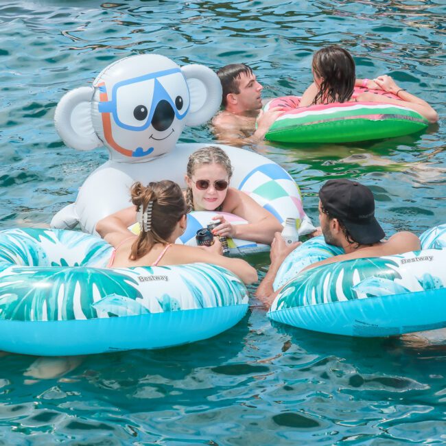 A group of people enjoying themselves in a body of water on inflatable floats. One float features a large koala design, while the others are decorated with blue and white patterns. Everyone appears relaxed and happy in the sun.